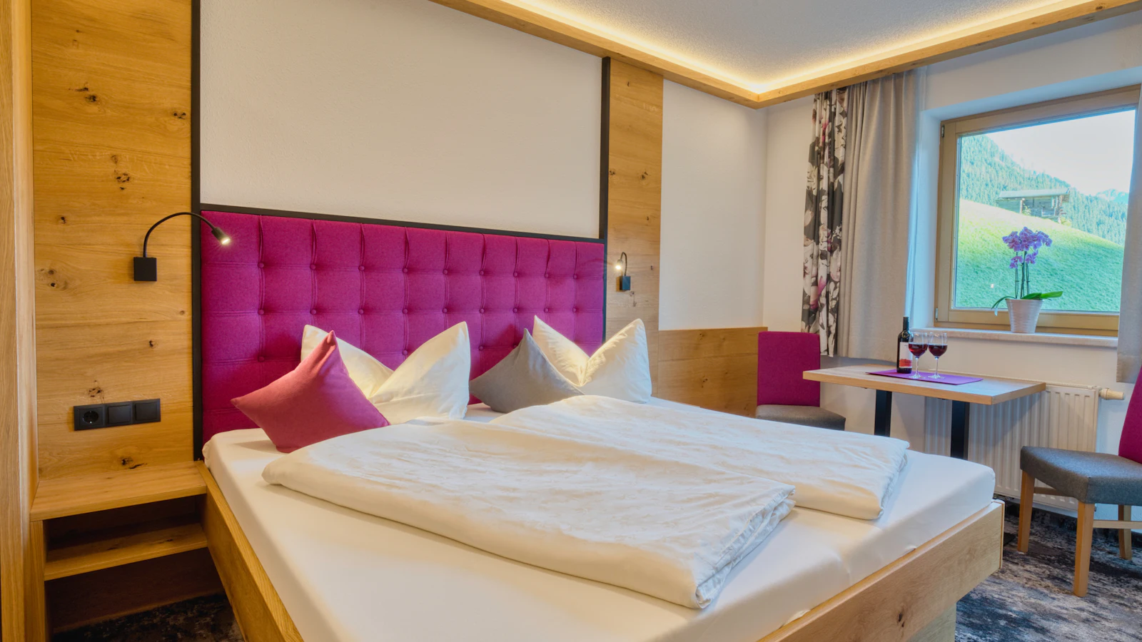 A guesthouse bedroom with a pink upholstered headboard, white bedding, a small table set with two glasses of red wine and a window looking out on green mountain meadows