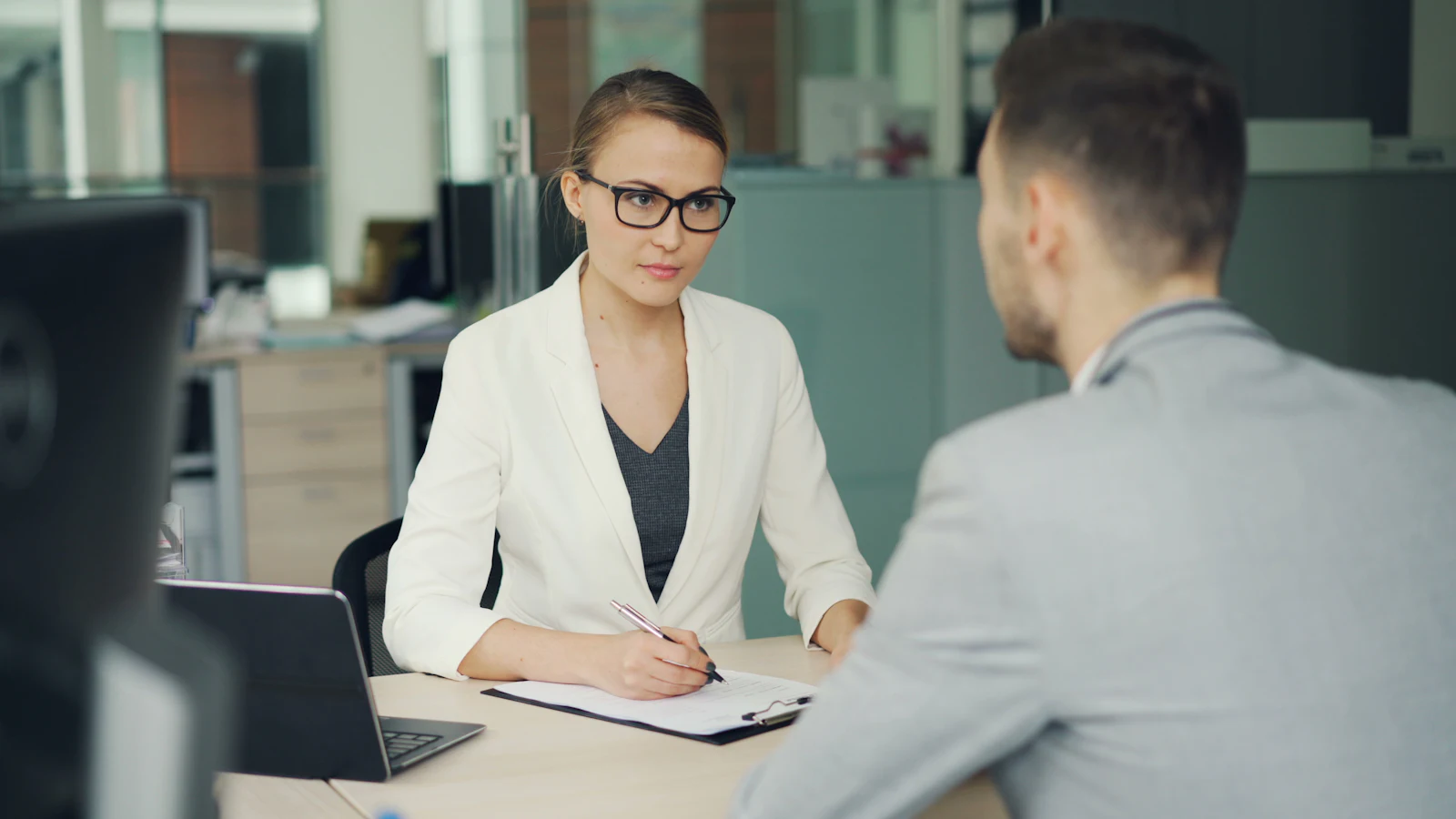 Recruiter in glasses taking notes on a clipboard while interviewing a candidate across an office desk